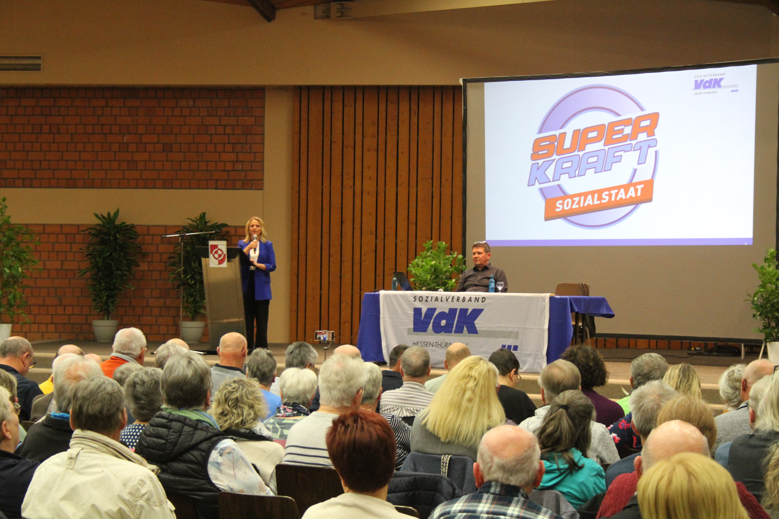Blick über Köpfe hinweg auf das Podium in der Halle, wo eine Frau und ein Mann an einem langen Tisch mit großem VdK-Logo sitzen und sprechen. 
