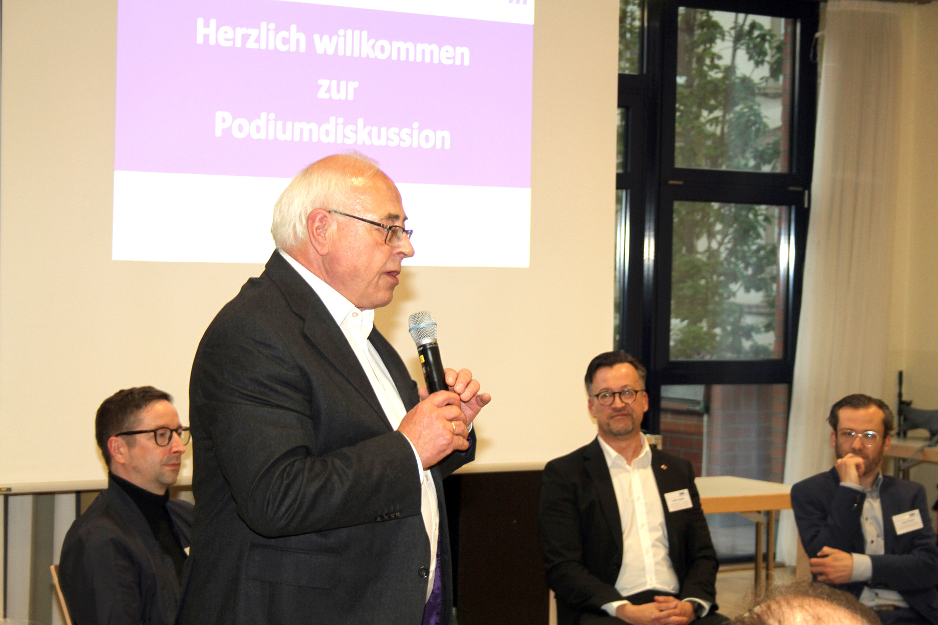 Ein Mann steht auf einer B&uuml;hne und spricht vor Publikum. Im Hintergrund sitzen die Teilnehmenden einer Podiumsdikussion.