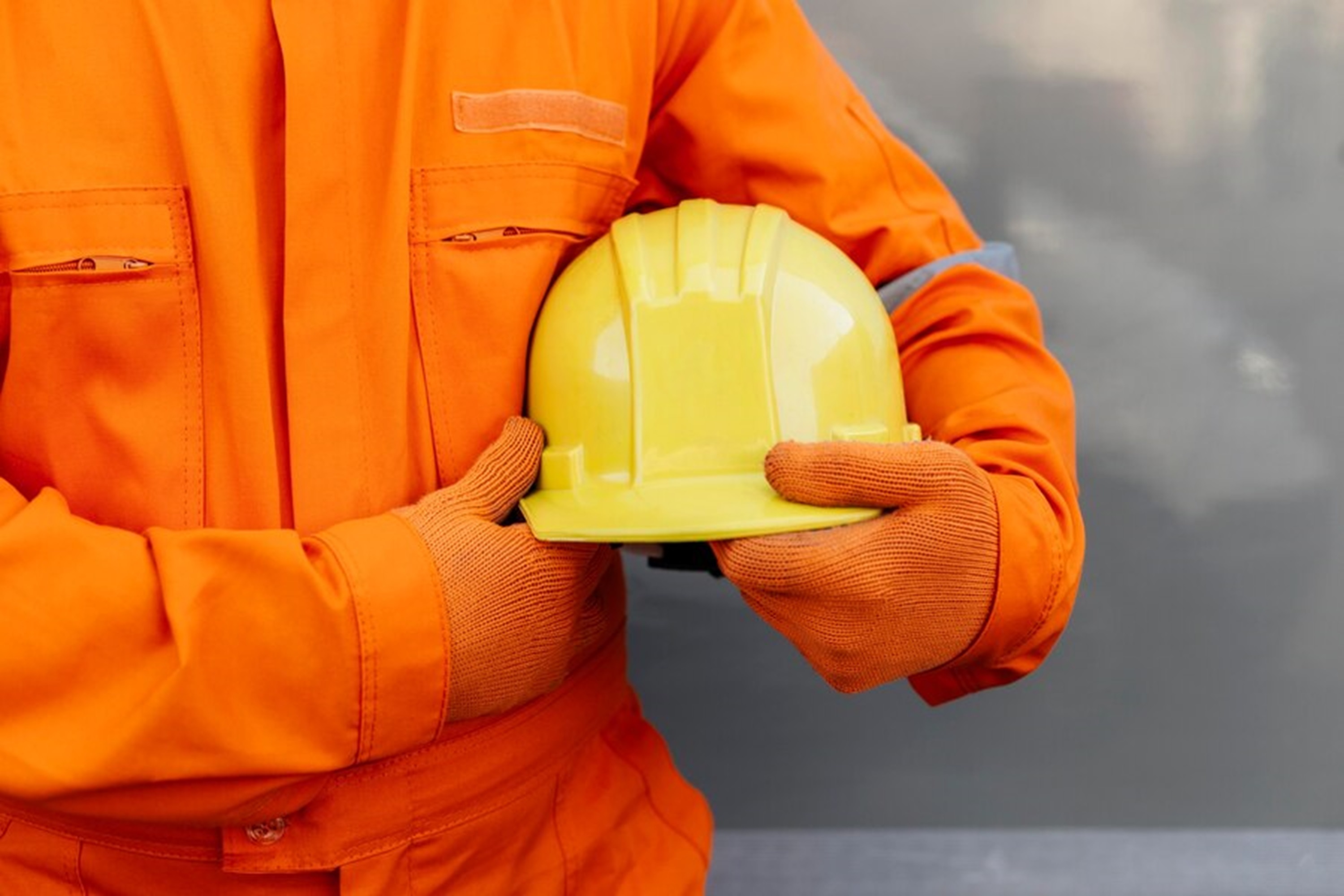 Ein Mensch in einem orangefarbenen Overall tr&auml;gt einen gelben Helm in den H&auml;nden