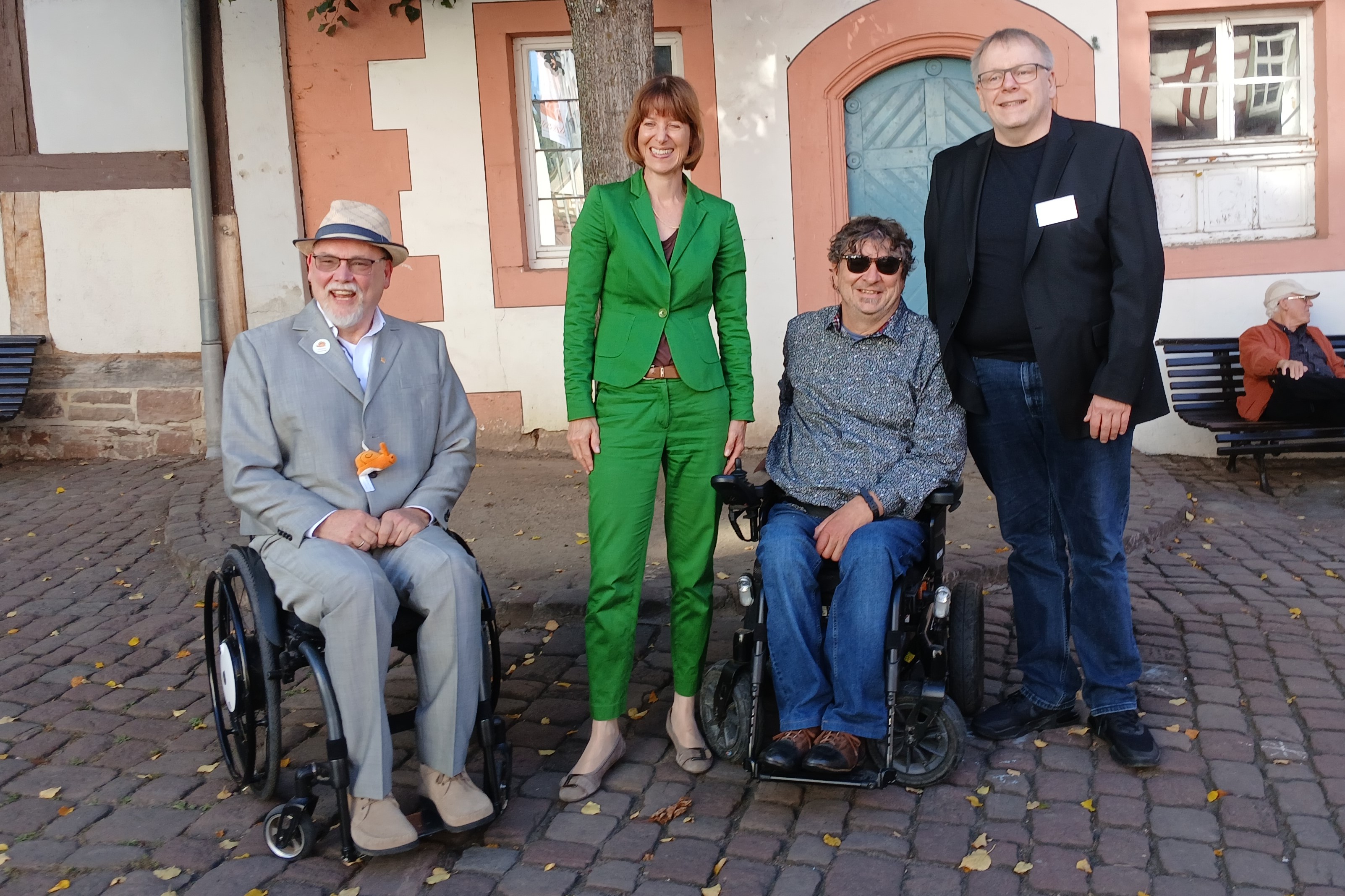 Zwei M&auml;nner im Rollstuhl stehen mit einer Frau im gr&uuml;nen Hosenanzug und einem Mann im schwarzen Jackett auf dem Kopfsteinpflaster vor einem Haus