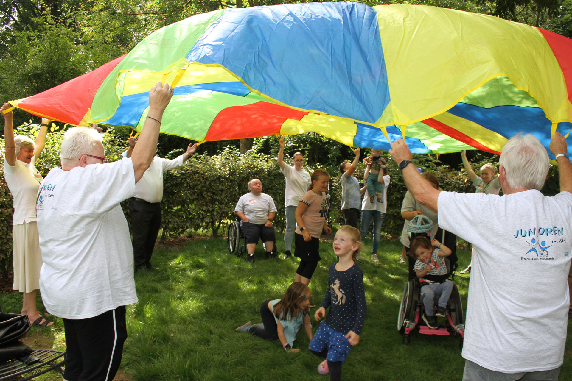Erwachsene und Kinder heben in einem Park ein gro&szlig;es, buntes Schwungtuch, w&auml;hrend Kinder darunter spielen.