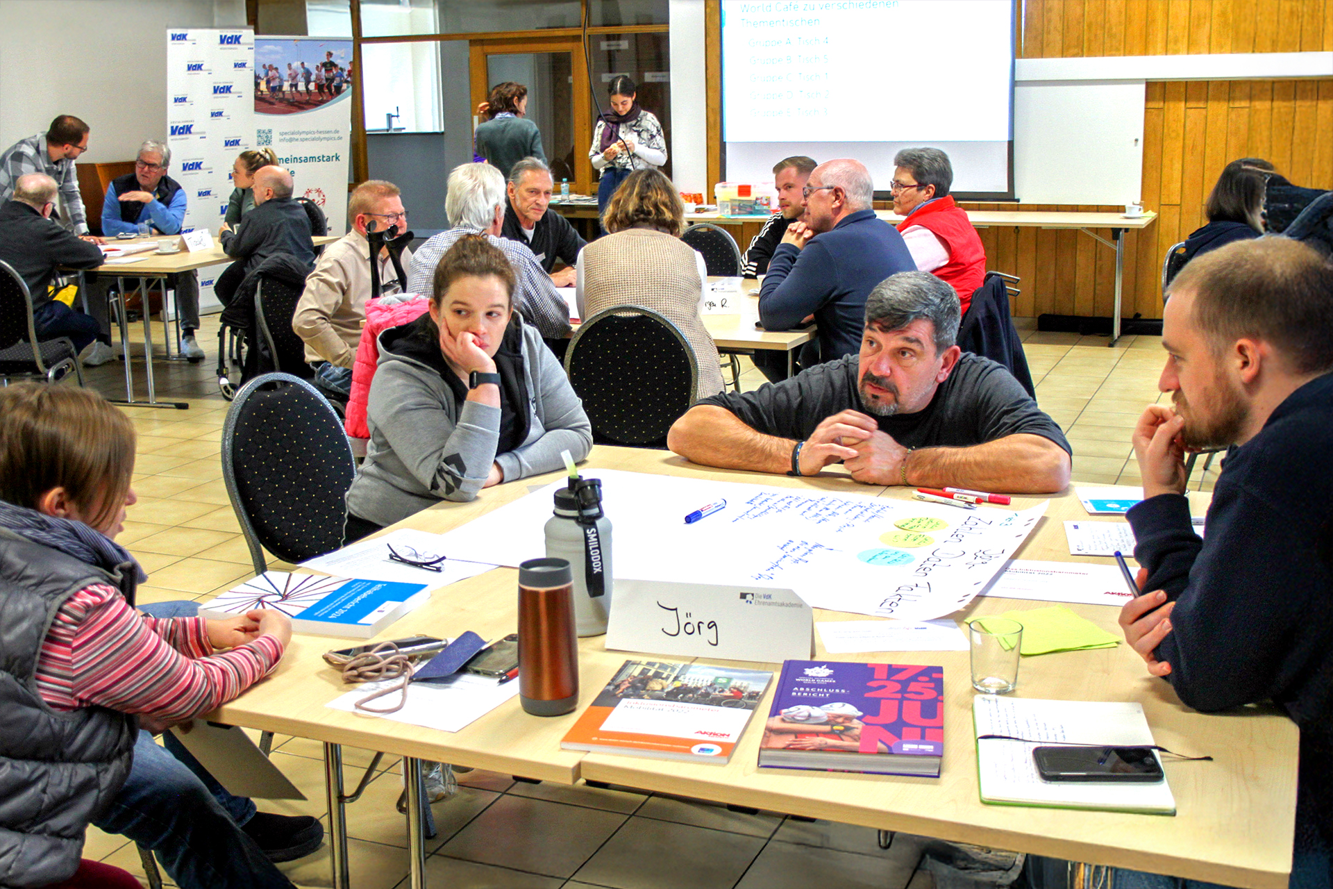Blick in einen gro&szlig;en Seminarraum. Im Hintergrund ist eine Leinwand heruntergezogen und es ist ein Roll-up des VdK-Hessen-Th&uuml;ringen aufgebaut. Die Menschen sitzen zu kleinen Gruppen an verschiedenen Tischen und unterhalten sich mit den anderen aus ihrer Gruppe.