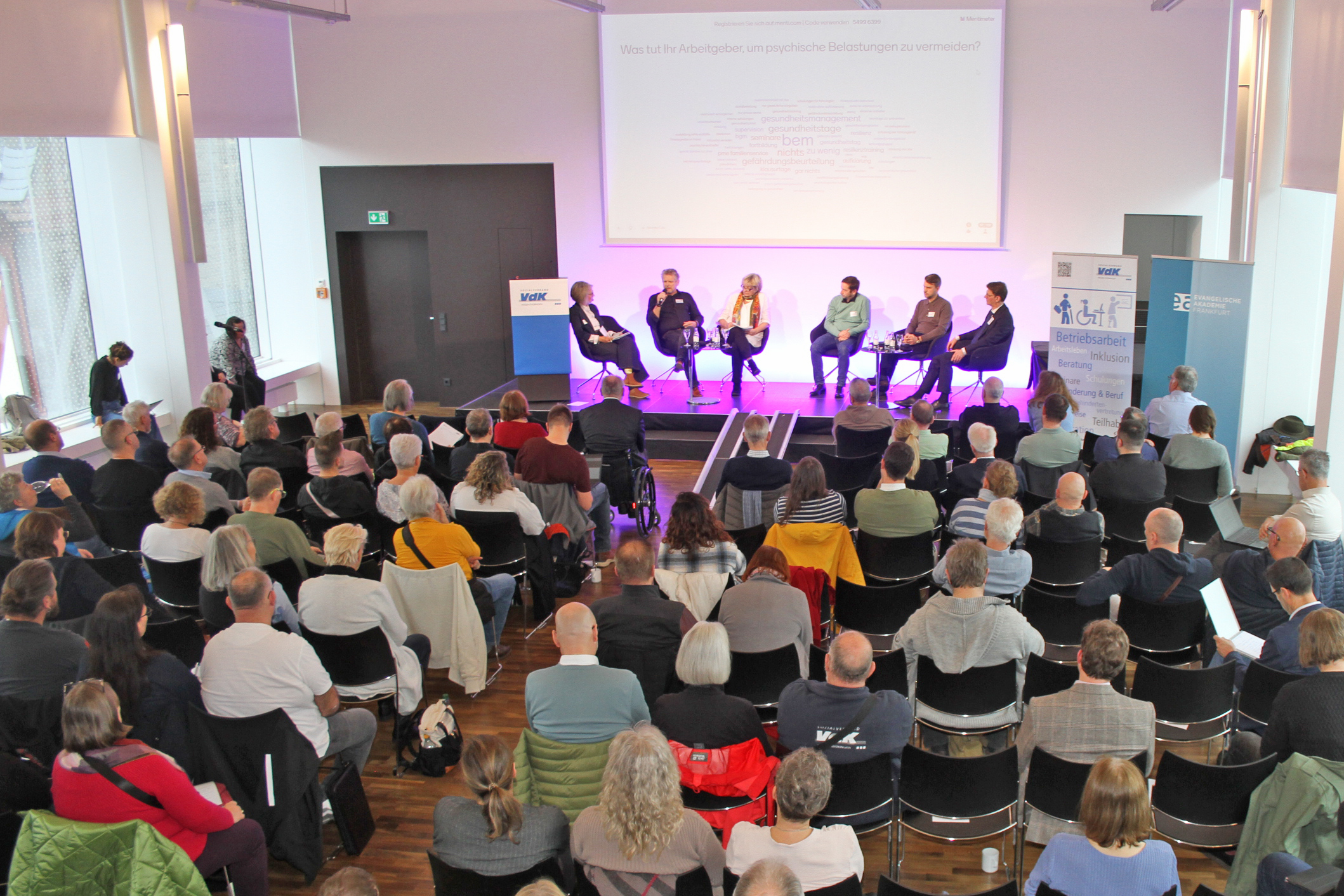 Ein Blick von oben in einen Vortragsraum. Der Raum ist gut gefüllt mit etwa 50 Gästen. Vorne ist eine kleine Bühne, auf der 6 Menschen sitzen, die an einer Podiumsdiskussion teilnehmen.