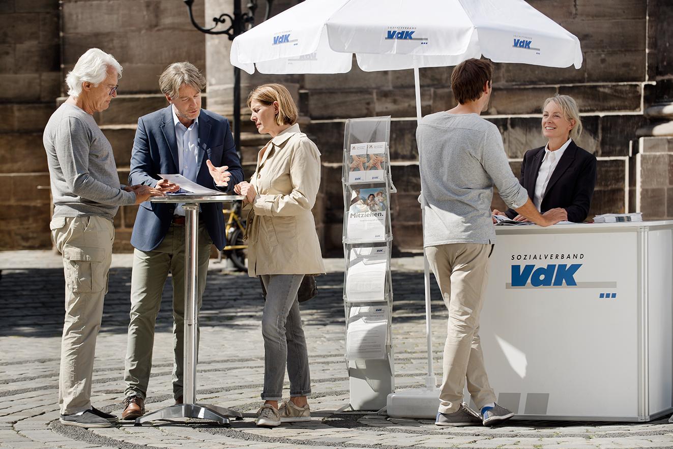 Ein VdK-Infostand in einer Fu&szlig;g&auml;ngerzone, eine Frau spricht mit einem Passanten, am Stehtisch daneben unterhalten sich Menschen. 