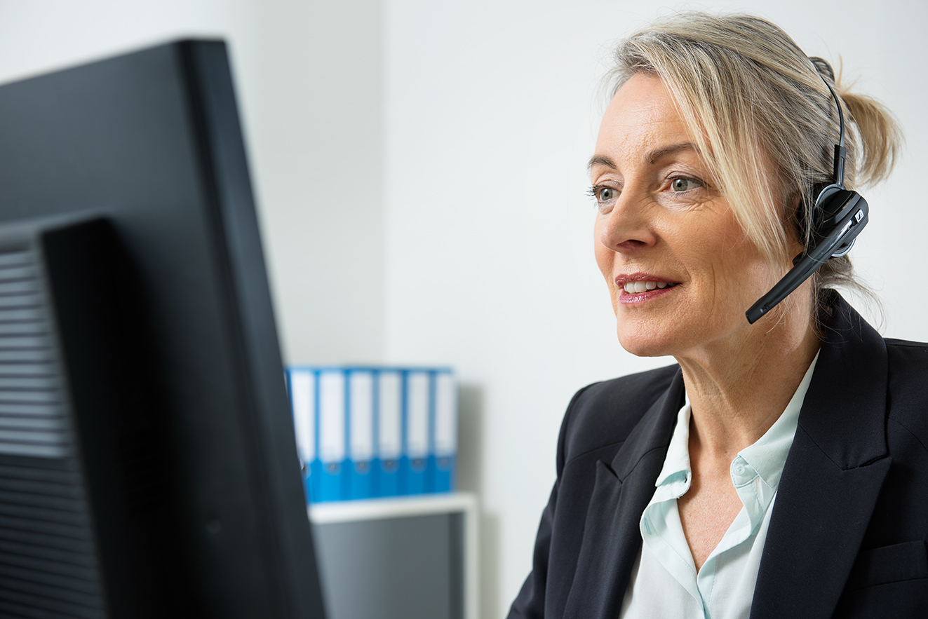 Eine Frau am Computer, sie telefoniert &uuml;ber ein Headset. Im Hintergrund sieht man Aktenordner.