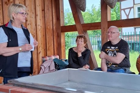 Andrea Fiedler steht links. 2 Personen sitzen im Bildhintergrund.