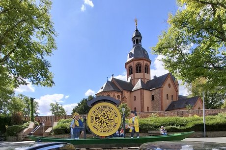 Im Hintergrund die Basilika, rechts und links eingerahmt von B&auml;umen. Davor steht, in einem flachen Kahn, ein rundes Gelbes Schild, etwas 2 Meter Hoch. Die Inschrift lautet "DER FISCHER ZVMT SIGEL IN SELGELSTATT 1546"