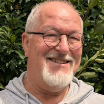 Portrait eines lächelnden, grauhaarigen Mannes, wenig Haare am Oberkopf, mit runder Brille und Ober-/Unterlippenbart.