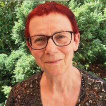 Lächelnde Frau in den Sechzigern, sehr kurze dunkelrote Haare, sie trägt eine größere, schwarzgeränderte Brille und ein gemustertes, dunkles Shirt, sie steht vor grünen Büschen und Bäumen.