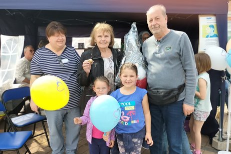 Das Bild zeigt die Hauptgewinner gemeinsam bei der Preis&uuml;bergabe am Stand des OV Nauheim. In der Mitte steht eine Familie, bestehend aus zwei Erwachsenen und zwei Kindern, die stolz ihren Gewinn pr&auml;sentieren. Die Kinder halten bunte Luftballons in den H&auml;nden und l&auml;cheln in die Kamera. Die erwachsene Frau in der Mitte h&auml;lt eine Gewinnkugel, w&auml;hrend neben ihr ein gro&szlig;er, verpackter Hauptpreis &uuml;berreicht wurde. Alle Beteiligten wirken sichtlich erfreut und zufrieden. Im Hintergrund ist der Veranstaltungsstand mit weiteren Besuchern und Dekorationen zu sehen, was die lebendige und gut besuchte Atmosph&auml;re unterstreicht. Insgesamt vermittelt das Bild einen gelungenen Moment der Veranstaltung, bei dem sich die Hauptgewinner &ndash; Familie XXX &ndash; &uuml;ber ihren 1. Preis freuen konnten.