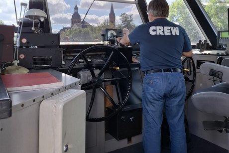 Blick durch die Br&uuml;cke auf die Altstadt. Der Kapit&auml;n ist von hinten zu sehen, er tr&auml;gt Jeans und ein T-Shirt mit der Aufschrift "CREW". Vor ihm sind zwei Steuerr&auml;der, die rechte Hand ist auf dem rechten Steuerrad, die linke Hand auf dem Geschwindigkeitshebel.