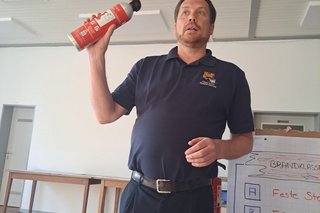 Benjamin Horst berichtete u.a. über die verschiedenen Feuerlöscher und ihre Funktion. Er steht im Raum und hält einen kleinen Feuerlöscher in der Hand.