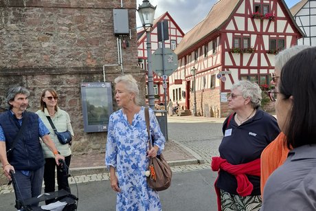 Auf der Stra&szlig;e in der Altstadt steht die Frendenf&uuml;hrerin in einem wei&szlig;-blauen Kleid und erz&auml;hlt etwas. Um sie herum stehen 5 Personen, einer mit Rollator.