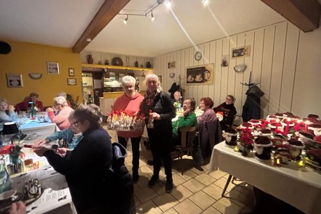 -Das Bild zeigt eine gem&uuml;tliche Zusammenkunft &auml;lterer Menschen in einem festlich dekorierten Raum, vermutlich eine Weihnachts- oder Adventsfeier. Was zu sehen ist: Viele Menschen sitzen an mehreren Tischen, trinken Kaffee oder Tee und unterhalten sich. Die Stimmung wirkt warm und gesellig. In der Raummitte stehen zwei Personen, die jeweils K&ouml;rbe mit kleinen Geschenken oder dekorierten Flaschen halten &ndash; m&ouml;glicherweise werden gerade Pr&auml;sente verteilt. Rechts steht ein gro&szlig;er Tisch voller weihnachtlicher Dekorationen, darunter kleine Weihnachtsm&auml;nner, Kerzen und festliche Arrangements. Die W&auml;nde sind gem&uuml;tlich dekoriert, u. a. mit Bildern, alten K&uuml;chenutensilien und einem Wandregal. Die Beleuchtung ist warm, was zur weihnachtlichen Atmosph&auml;re beitr&auml;gt. Insgesamt wirkt es wie eine liebevoll organisierte Adventsfeier in einem Gemeinschaftsraum oder Vereinslokal.