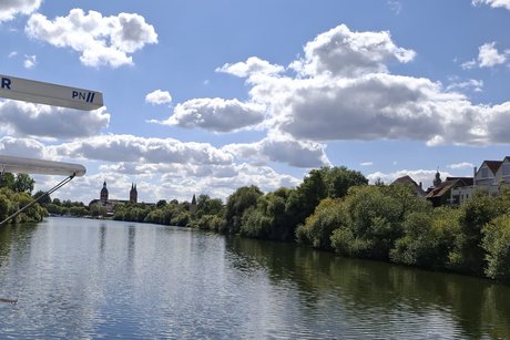 Blick vom Schiff nach vorn. In der Ferne sind zwei T&uuml;rme von der Altstadtzu sehen. Das Ufer ist rechts und links mit mit B&auml;umen und B&uuml;schen dicht bewachsen. Dar&uuml;ber ein blauer Himmel mit wei&szlig;en Wolken. Alles spiegelt sich im Wasser des Flusses.