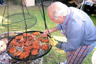 Ein Schwenkgrill auf einem Rasen. Ein älterer Herr mit Schürze hält ihn zur Seite und wendet die Fleischstücke