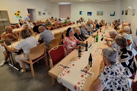 Ein kleiner Saal mit G&auml;sten an drei Tischreihen, einige St&uuml;hle sind noch leer.