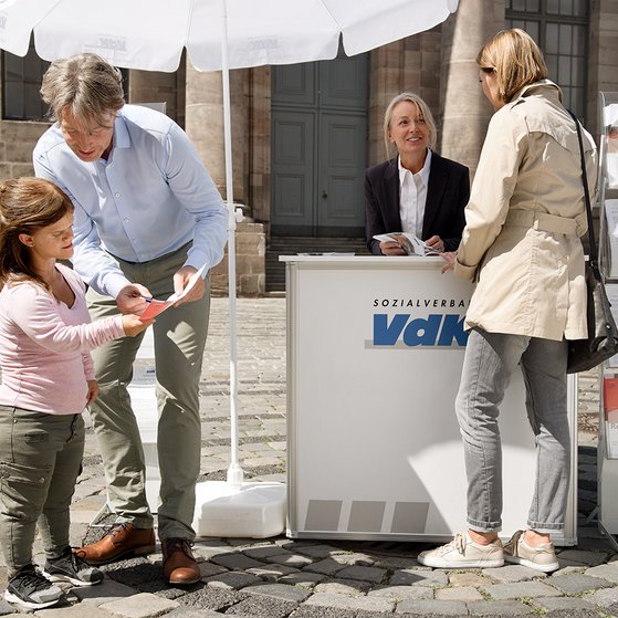 VdK-Infostand Ein VdK-Infostand mit einer Mitarbeiterin, davor verschiedene Menschen, die sich informieren wollen.