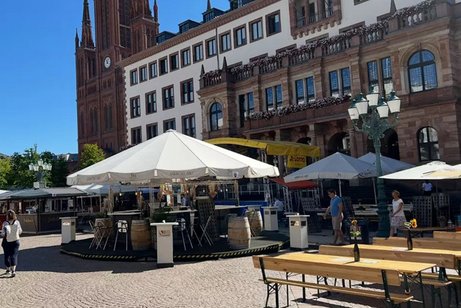 Gebäudeansicht des Wiesbadener Rathauses, im Vordergrund Außengastronomie mit Biertischen und Sonnenschirmen