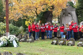 Die beiden Kränze liegen am Fuße eines großen Holzkreuzes. Dahinter sipelt die Bläsergruppe in roten Jacken.