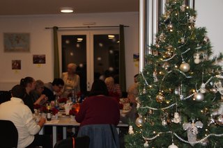 Ein stimmungsvolles Bild mit einem festlich geschmückten Weihnachtsbaum im Vordergrund rechts. Auf der linken Bildseite sieht man - etwas dunkler und im Hintergrund - einen Tisch um den herum die Besucher sitzen.