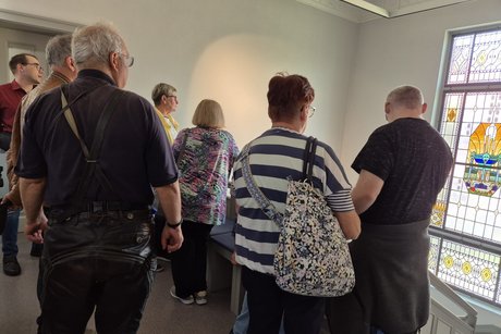 Die Gäste schauen auf ein gestaltetes Buntglasfenster. Im Hintergrund steht der Museumsleiter.