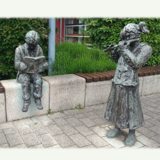 Hänsel und Gretel heißen die beiden Statuen vor der Bücherei in Liederbach. zu sehen sind 2 statuen die vor der bücherei liederbach aufgestellt sind. ein lesender junge sitzt auf der steinmauer vor der bücherei. das mädchen steht und spielt flöte