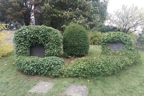 Das Bild zeigt das Ehrenmal für die Opfer der beiden Weltkriege. Auf den Gedenksteinen stehen die Namen der Opfer auf einer Metallplatte. Der Gedenkstein ist mit Efeu überwachsen