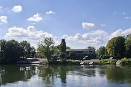 Unter einem blauen Himmel mit kleinen, wei&szlig;en Wolken liegt - umgeben von B&auml;umen - die alte F&auml;hranlegestelle. Das Ufer mit den B&auml;umen spiegelt sich im Wasser.