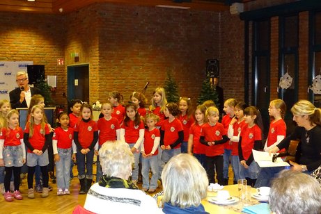Auf dem Foto sind die Mini Voices und ihre Leiterin Martine Gillner (rechts) sowie der Moderator der Weihnachtsfeier, Holger Kintscher (links), zu sehen