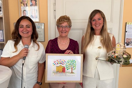 KiTa-Leiterin Barbara Kus (links) und Doro Kissnè-Szabo (rechts) übergaben der VdK-Vorsitzenden Ruth Deiseroth (Mitte) ein von Kindern der KiTa selbst gemaltes Bild und ein Haus mit 40 guten Wünschen für die Begegnungsstätte.en KiTa-Leiterin Barbara Kus (links) und Doro Kissnè-Szabo (rechts) übergaben der VdK-Vorsitzenden Ruth Deiseroth (Mitte) ein von Kindern der KiTa selbst gemaltes Bild und ein Haus mit 40 guten Wünschen für die Begegnungsstätte.