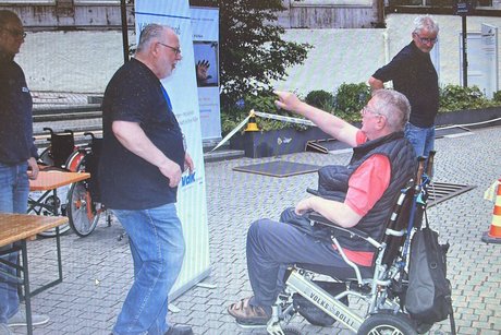 Ein Mann sitzt im Rollstuhl. Mehrere Personen stehen dabei.