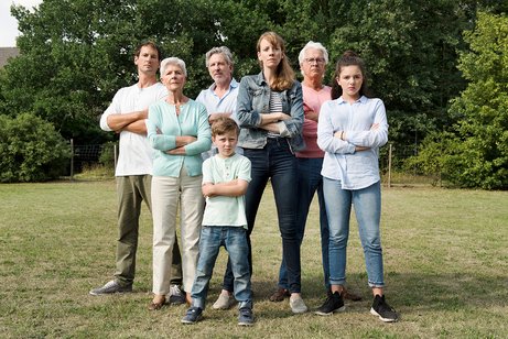 Eine Gruppe von Erwachsenen und Kindern steht als Gruppe dicht beeinander, sie blicken ernst, haben die Arme ablehnend verschr&auml;nkt. 