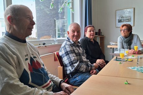 ein Tisch im Raum, auf einer Seite sitzen 4 Personen die sich unterhalten oder l&auml;chelnd in die Kamera schauen