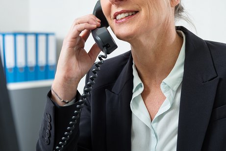 Eine Frau am Computer, sie telefoniert. Im Hintergrund sieht man Aktenordner.