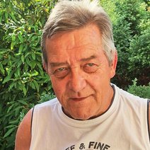 Brustbild eines Mannes, Mitte 60, kurze, angegraute Haare, er trägt ein weißes Achselshirt und steht vor grünen Büschen und Bäumen.