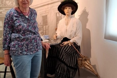 Im Hintergrund sitzt eine Puppe im historischen Kostüm. Links steht eine Frau.