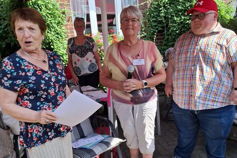 Drei Personen stehen im Vordergrund. Eine weitere Frau steht im Hintergrund.