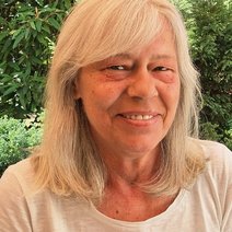 Eine lächelnde Frau, Mitte 60, blonde Haare, schulterlang und glatt, mit Seitenscheitel rechts, sie trägt ein weißes Shirt und steht vor grünen Büschen und Bäumen.