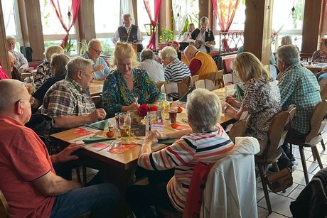 Das Bild zeigt einen lebhaften Gastraum mit zahlreichen Gästen an mehreren Holztischen. Die Szene wirkt wie eine gemeinsame Veranstaltung, ein Vereins- oder Seniorentreffen in einem Restaurant oder Lokal.
