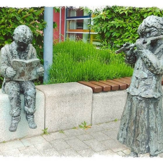 Auf dem Bild sind 2 Statuen zu sehen, die vor der Bücherei stehen. Ein lesender Junge und ein Mädchen das Flöte spielt sind zu sehen.