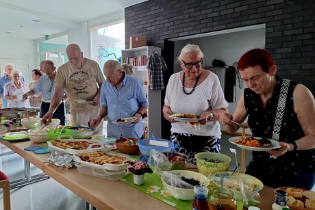 Eine Tischreihe mit Salaten Grillgut und anderen Leckereien. Davor eine kleine Menschenschlange.
