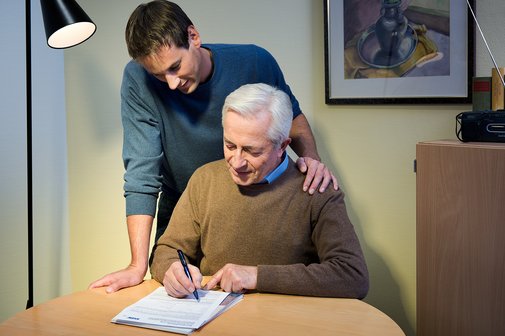 VdK-Beitrittserklärung Ein älterer Mann sitzt an einem Küchentisch und füllt eine VdK-Beitrittserklärung aus, ein jüngerer Mann blickt ihm über die Schulter.