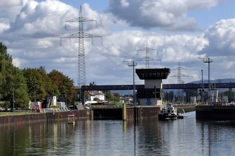 Blick auf die Schleusentore vom Schiff aus. Das rechte Schleusentor ist offen, ein kleines Schiff f&auml;hrt da heraus. Dar&uuml;ber sieht man Hochspannungsmasten und wei&szlig;e Wolken.