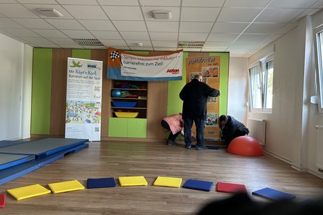 Das Bild zeigt die Vorbereitung f&uuml;r den Parcours, einen gro&szlig;en Raum vorne mit Sitzkissen im Halbkreis, hinten einen Mann der Rollups aufbaut