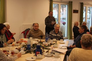 Um einen weihnachtlich gedekten Tisch, mit Kaffee und Gebäck, sitzen 7 Menschen. Dahinter steht Herr Hanke und lies aus einem kleinen Buch ein Gedicht vor.