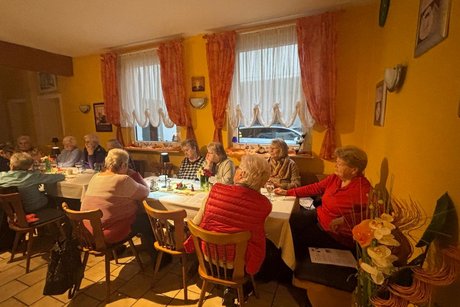 .Das Bild zeigt eine gem&uuml;tliche Zusammenkunft &auml;lterer Menschen in einem festlich dekorierten Raum, vermutlich eine Weihnachts- oder Adventsfeier. Was zu sehen ist: Viele Menschen sitzen an mehreren Tischen, trinken Kaffee oder Tee und unterhalten sich. Die Stimmung wirkt warm und gesellig. In der Raummitte stehen zwei Personen, die jeweils K&ouml;rbe mit kleinen Geschenken oder dekorierten Flaschen halten &ndash; m&ouml;glicherweise werden gerade Pr&auml;sente verteilt. Rechts steht ein gro&szlig;er Tisch voller weihnachtlicher Dekorationen, darunter kleine Weihnachtsm&auml;nner, Kerzen und festliche Arrangements. Die W&auml;nde sind gem&uuml;tlich dekoriert, u. a. mit Bildern, alten K&uuml;chenutensilien und einem Wandregal. Die Beleuchtung ist warm, was zur weihnachtlichen Atmosph&auml;re beitr&auml;gt. Insgesamt wirkt es wie eine liebevoll organisierte Adventsfeier in einem Gemeinschaftsraum oder Vereinslokal.