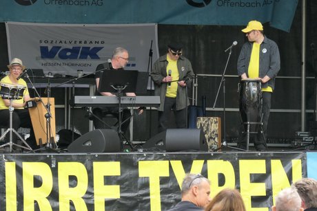 Auf der Bühne ist die Band "Irre Typen". 4 Männer in dunkelgrauer Kleidung mit gelben Hemden. Sie spielen Keyboard, Mundharmonika und Perkussion.