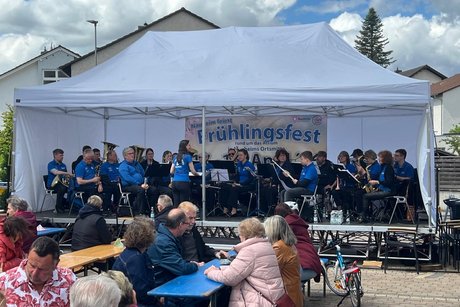 Das Bild zeigt eine lebendige Szene des Fr&uuml;hlingsfestes unter freiem Himmel. Im Mittelpunkt steht eine B&uuml;hne mit einem gro&szlig;en wei&szlig;en Zelt, unter dem ein Blasorchester sitzt. Die Musikerinnen und Musiker tragen einheitliche blaue Oberteile und spielen verschiedene Instrumente, w&auml;hrend eine Dirigentin vor ihnen steht und das Ensemble leitet. Im Hintergrund h&auml;ngt ein Banner mit der Aufschrift &bdquo;Fr&uuml;hlingsfest&ldquo;, das auf die Veranstaltung hinweist. Vor der B&uuml;hne sitzen zahlreiche Besucherinnen und Besucher an Bierzeltgarnituren, unterhalten sich und genie&szlig;en die Atmosph&auml;re. Die Stimmung wirkt entspannt und gesellig. Am Rand ist ein abgestelltes Fahrrad zu sehen, und im Hintergrund stehen Wohnh&auml;user sowie ein hoher Baum unter einem teils bew&ouml;lkten Himmel. Insgesamt vermittelt das Bild den Eindruck eines gut besuchten, gem&uuml;tlichen Dorffestes mit musikalischer Begleitung.