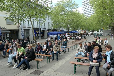 Auf dem Platz sind Holzbänke aufgestellt. Im vorderen Bereich sitzen Zuschauer.